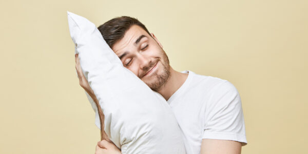 Horizontal image of handsome cute young man with bristle posing with head on white soft pillow sleeping peacefully and smiling, seeling good dream. Attractive guy napping after hard working day